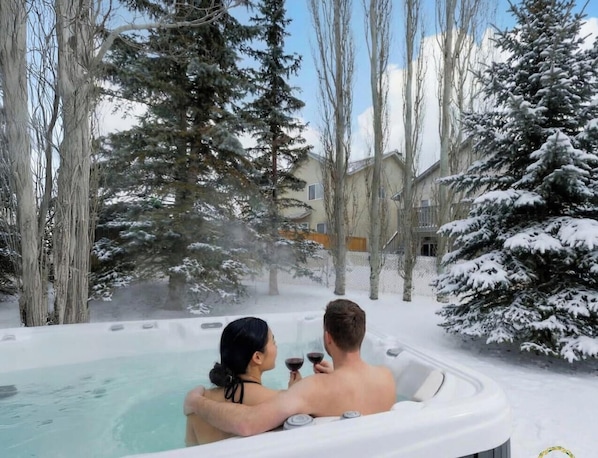 A hot tub with two people, surrounded by snow-covered trees and a building in the background.