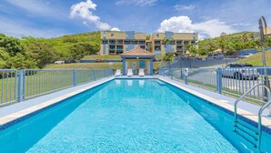 Piscina retangular com borda azulejada, rodeada por uma cerca de metal e um pequeno pavilhão com cadeiras no centro.