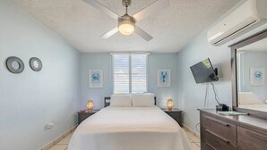 Quarto com cama, ventilador de teto, televisão e cômoda.