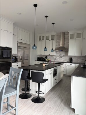 Una cocina moderna con armarios blancos, una isla central con taburetes de bar, electrodomésticos de acero inoxidable e iluminación colgante.