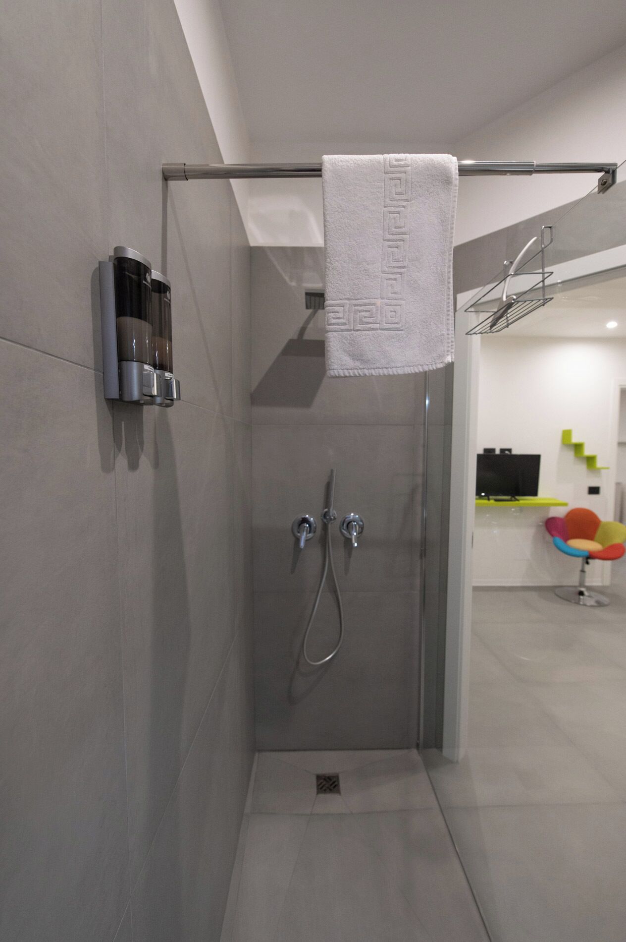 Una doccia moderna con un asciugamano appeso a un porta asciugamani, un dispenser di sapone a parete e uno scarico ben visibile.
