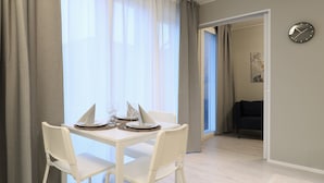 A dining area with a white table set for two, two white chairs, and a window with sheer curtains. - Forenom Serviced Apts Espoo Perusmäki 2 (Espoo)