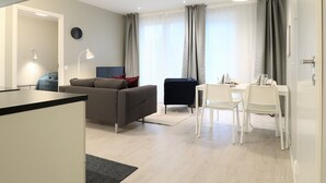 A modern living room with a grey sofa, a small black chair, and a dining table with white chairs. There is a flat-screen TV on a stand, a lamp, and a clock on the wall. - Forenom Serviced Apts Espoo Perusmäki 2 (Espoo)