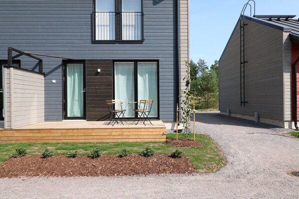 Una moderna casa a due piani con una veranda in legno, un piccolo giardino e un vialetto sterrato.