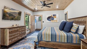 A bedroom with a wooden ceiling, a bed with a striped bedspread, a dresser, and a large painting on the wall.