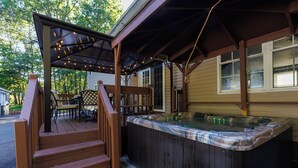 Una terraza de madera con jacuzzi, mobiliario exterior y una zona cubierta con guirnaldas de luces.