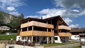 A multi-story wooden building with balconies, set against a mountainous backdrop. - New vacation apartment in the middle of the SkiArena Andermatt Sedrun with indoor golf course (Camischolas)