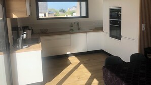 A modern kitchen with a window, white cabinets, and a dishwasher. - Seaside villa PMR standard Portsall (Ploudalmézeau)