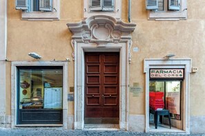 A building facade with a central wooden door, a shop window with a sign reading "FARMACIA DEL CORSO", and a small table with boxes outside.