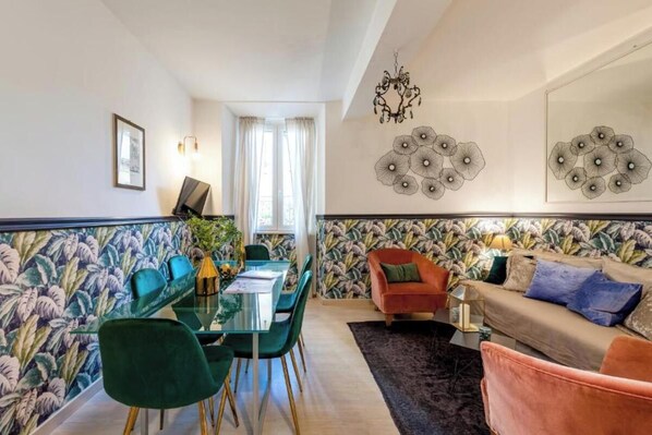 A modern living room with a glass dining table, green chairs, a red sofa, and a patterned wallpaper.