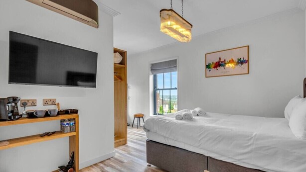 A modern hotel room with a large flat-screen TV, a wooden shelf with amenities, a bed with white linens, a colorful cityscape painting, and a view through a large window.