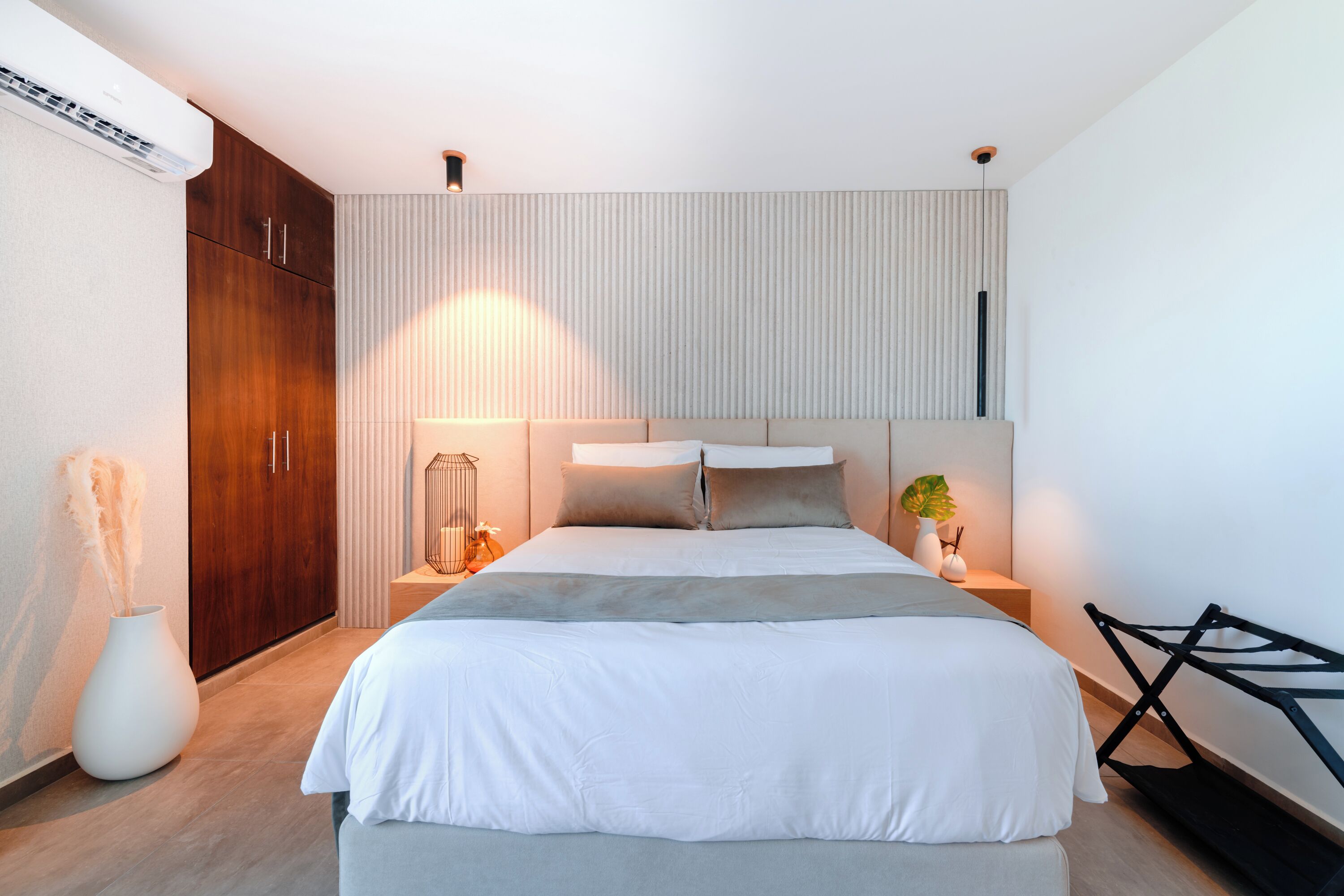 A modern bedroom with a large bed, wooden wardrobe, bedside tables with lamps, and a white wall with vertical blinds.