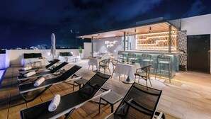 A rooftop bar with lounge chairs and tables, a patterned wall, and a view of the city skyline at night. - Areia Boutique Hotel - Puerto Morelos (Puerto Morelos)