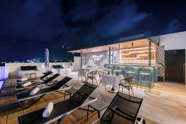 A rooftop bar with lounge chairs and tables, a patterned wall, and a view of the city skyline at night.