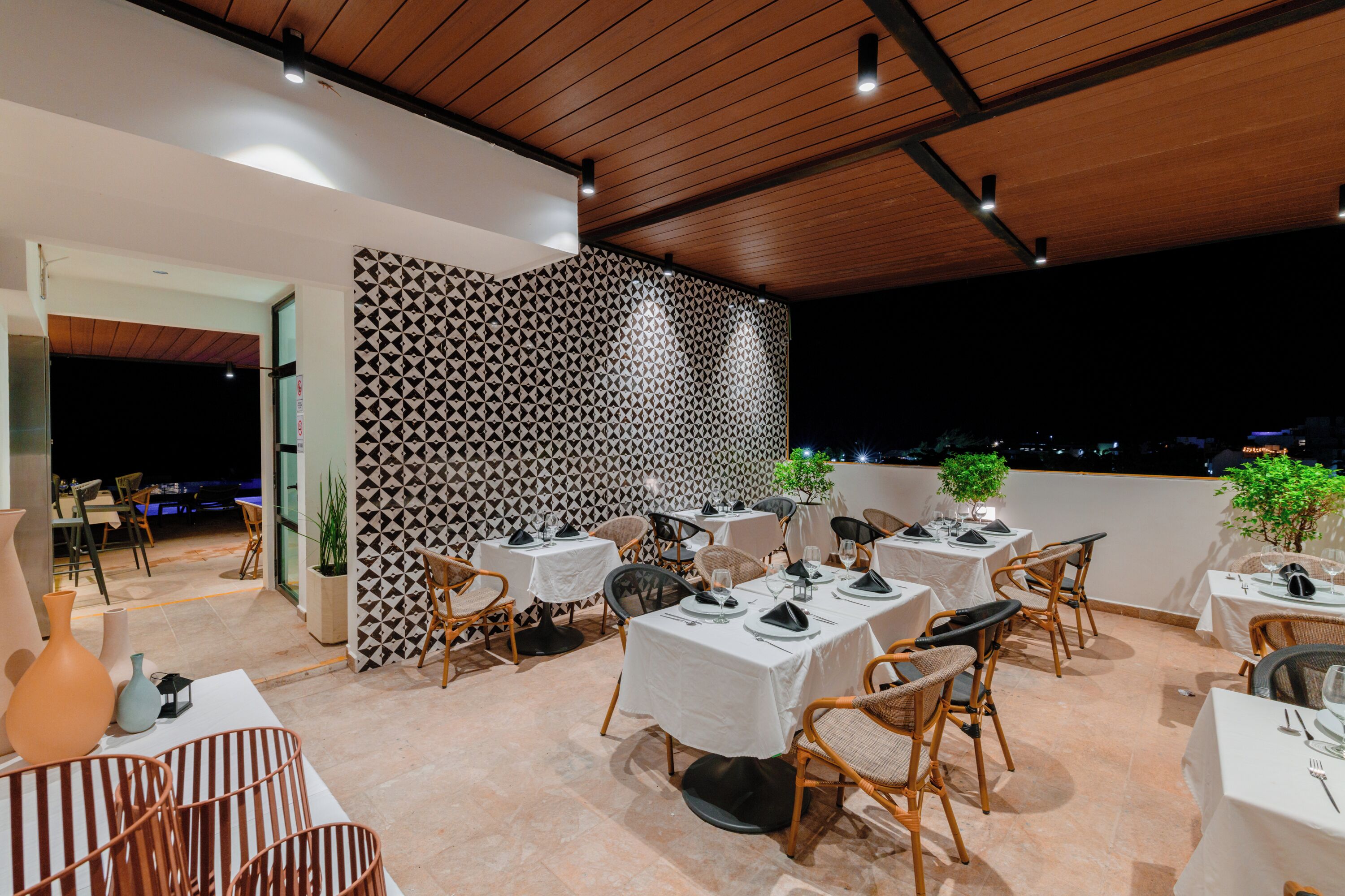 Un área de comedor al aire libre con mesas y sillas, un muro con diseño y vista a la ciudad de noche.