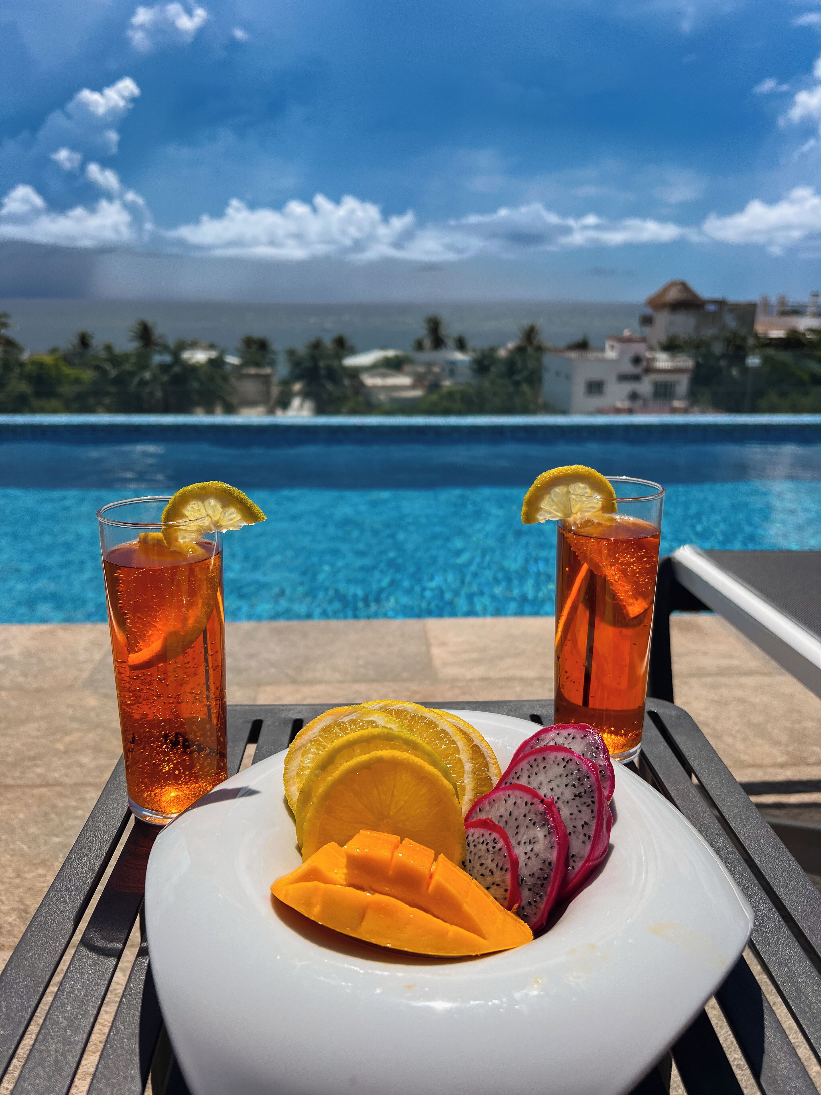 Una mesa con dos vasos de té helado, un plato con mango y pitahaya, y vista a una piscina y al océano.