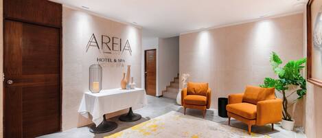 A modern hotel lobby with a reception desk, two armchairs, a potted plant, and a staircase in the background.