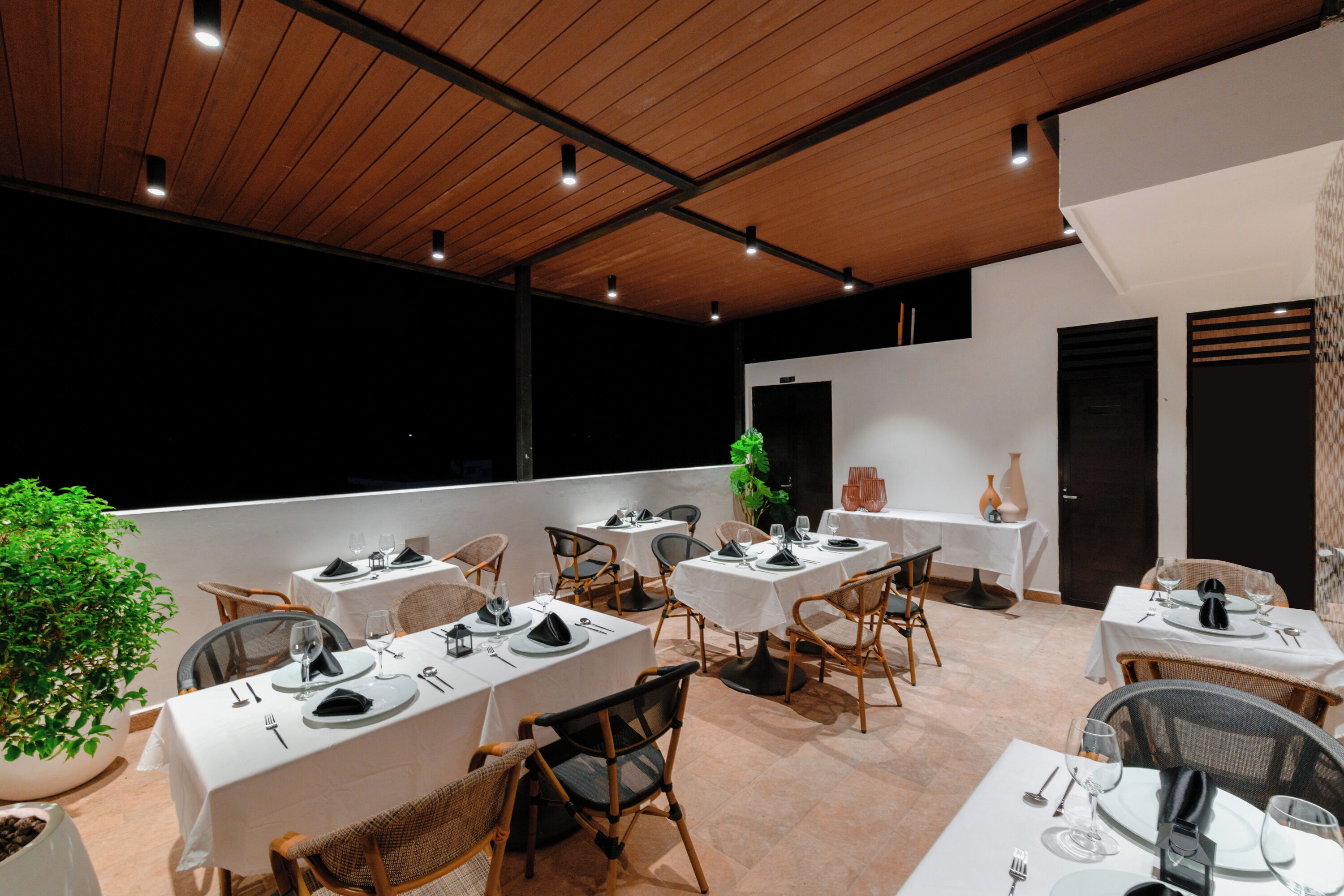 A restaurant with multiple tables set for dining, featuring a mix of chairs and tables, a potted plant, and a view of the night sky.