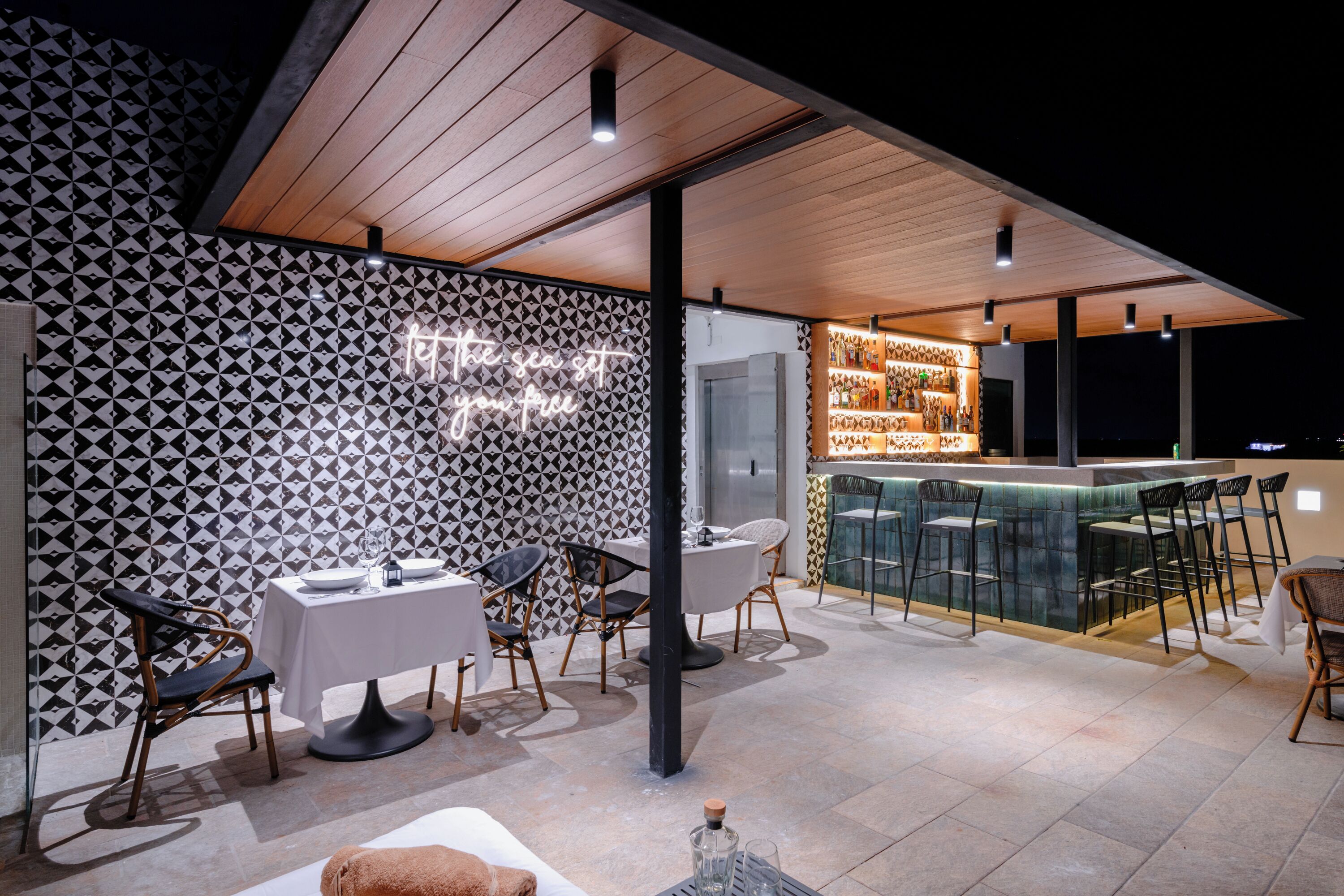 A modern outdoor dining area with a bar, featuring geometric patterned walls, wooden ceiling, and a view of the sea.