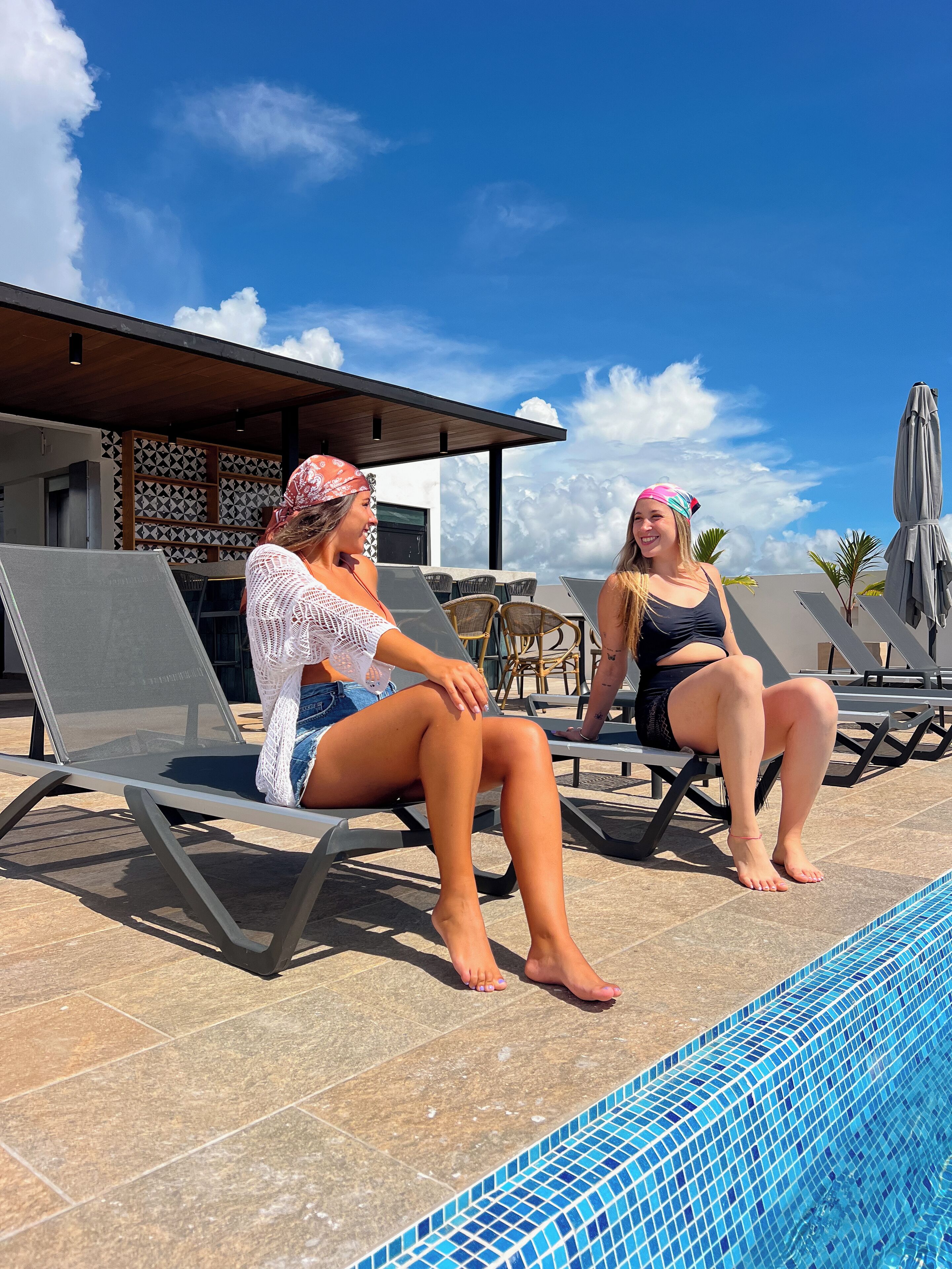 Two people sitting on lounge chairs by a pool with a tiled deck and a building with patterned walls in the background.