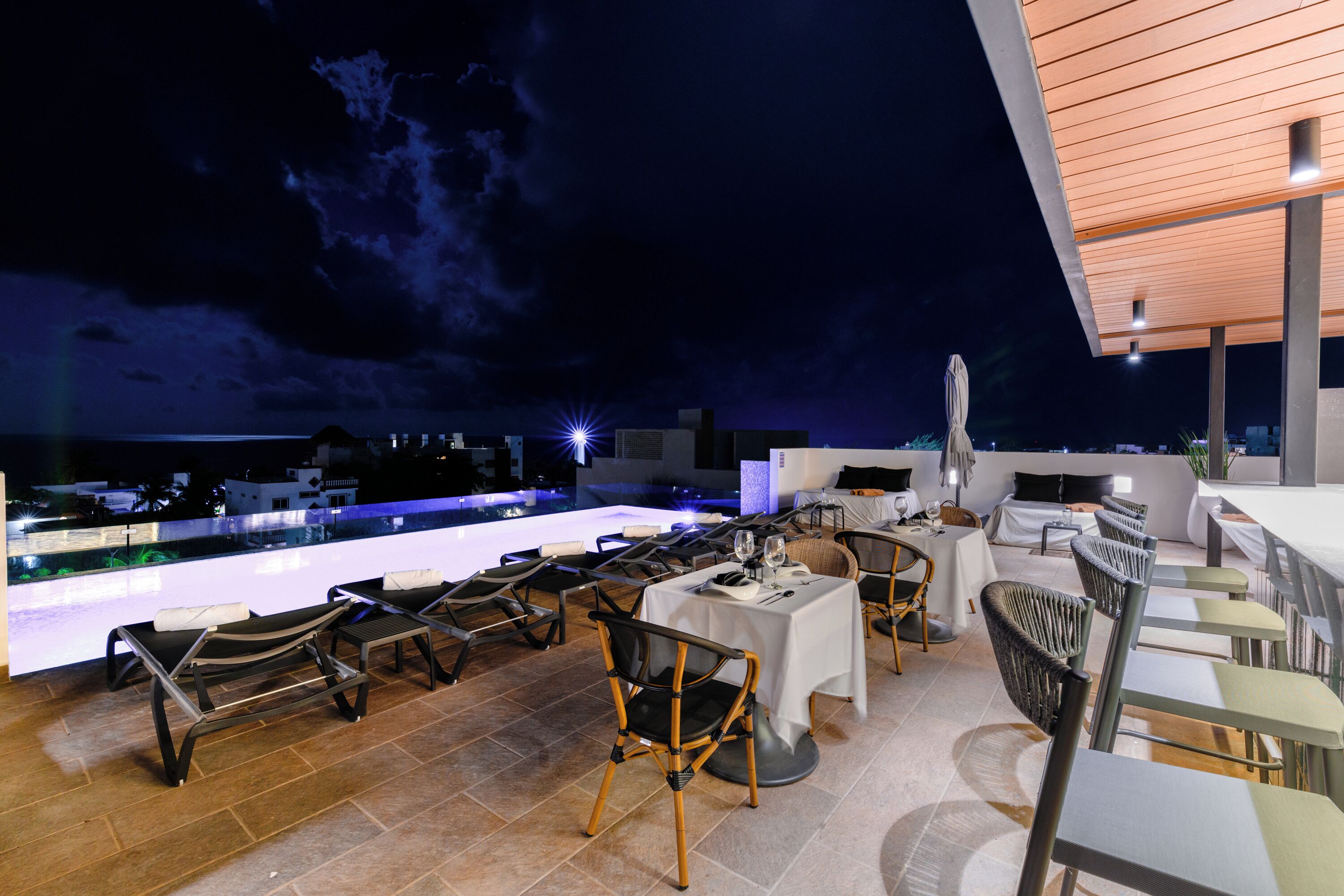 Ein Außenbereich zum Essen mit Tischen und Stühlen, ein Pool und Blick auf die Skyline der Stadt bei Nacht.
