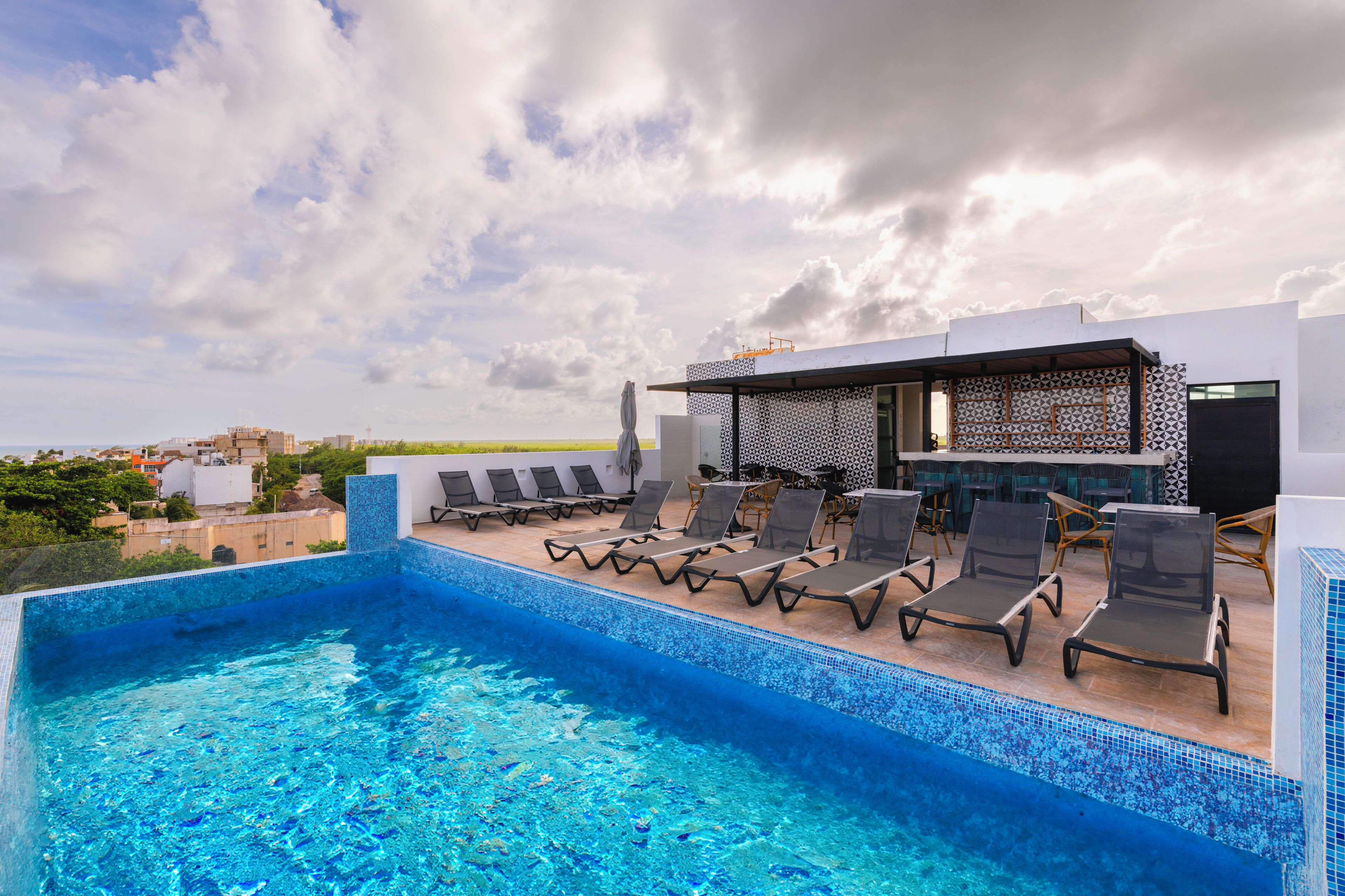 A rooftop pool with lounge chairs and a bar area, overlooking a cityscape.