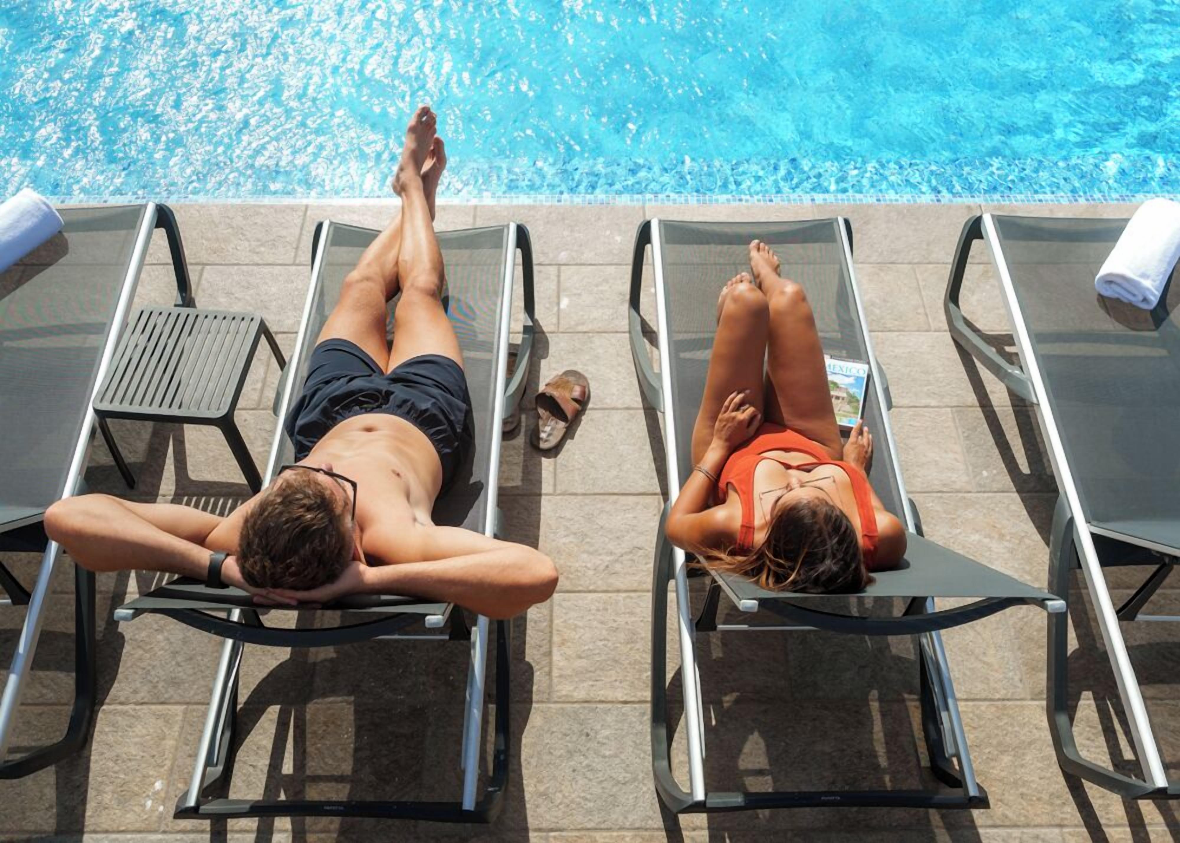 Two lounge chairs by a pool with a tiled floor and a small table.