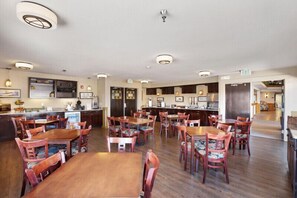 Un'area ristorazione di un hotel con diversi tavoli e sedie in legno, un angolo bar e una cucina sullo sfondo.