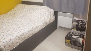 Ein kleines Zimmer mit einem Einzelbett, einem grauen Vorhang, einer Bettdecke mit Muster, einem weißen Heizkörper und einem kleinen Regal mit Gegenständen.