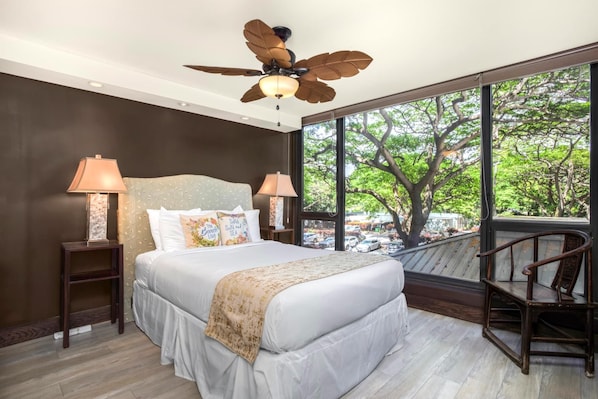 Une chambre à coucher avec un grand lit, deux lampes de chevet, un ventilateur de plafond et une vue sur les arbres à travers de grandes fenêtres.