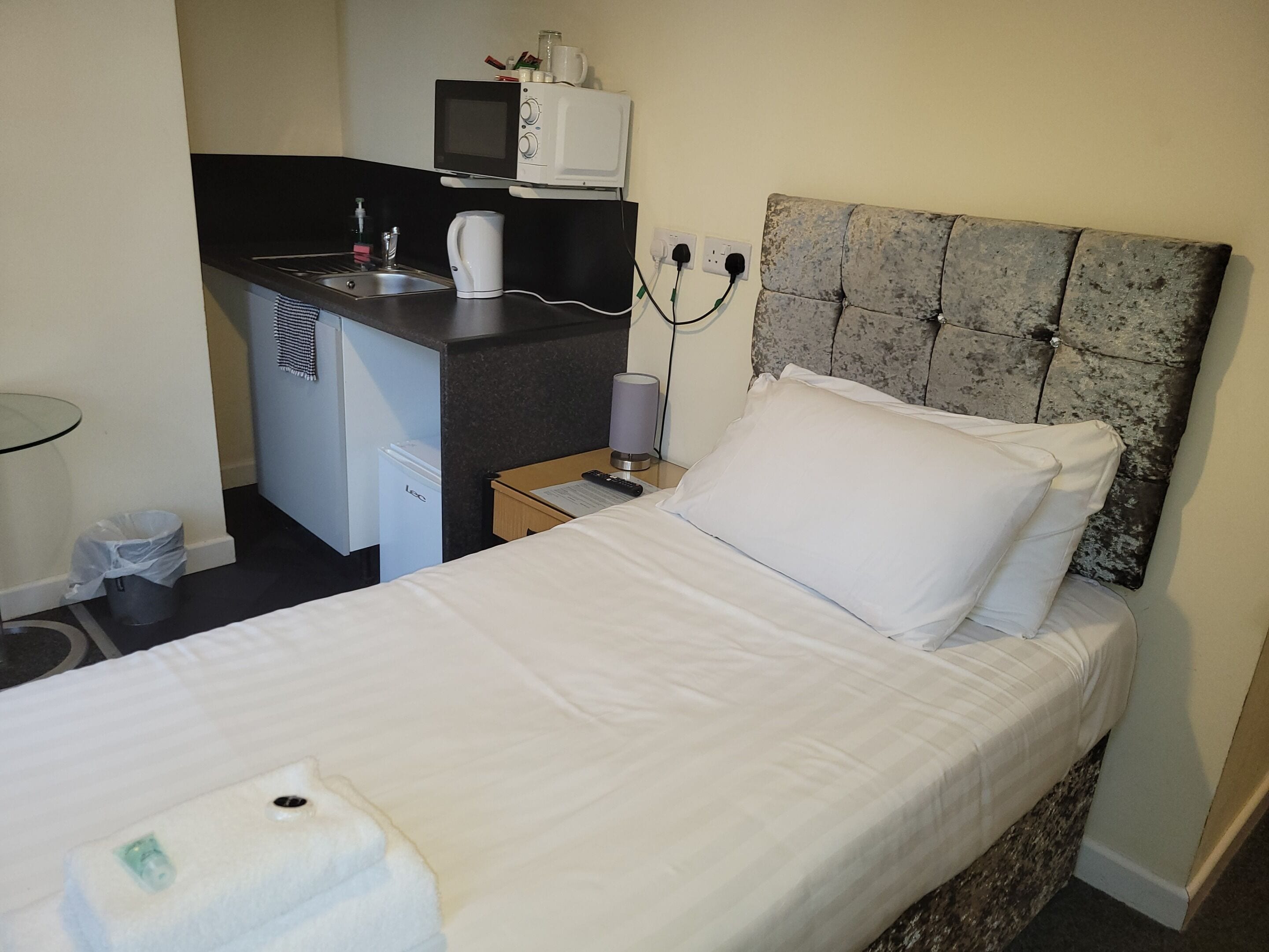 Hotelkamer met een bed, een klein kitchenette, een prullenbak en een nachtkastje met een lamp.