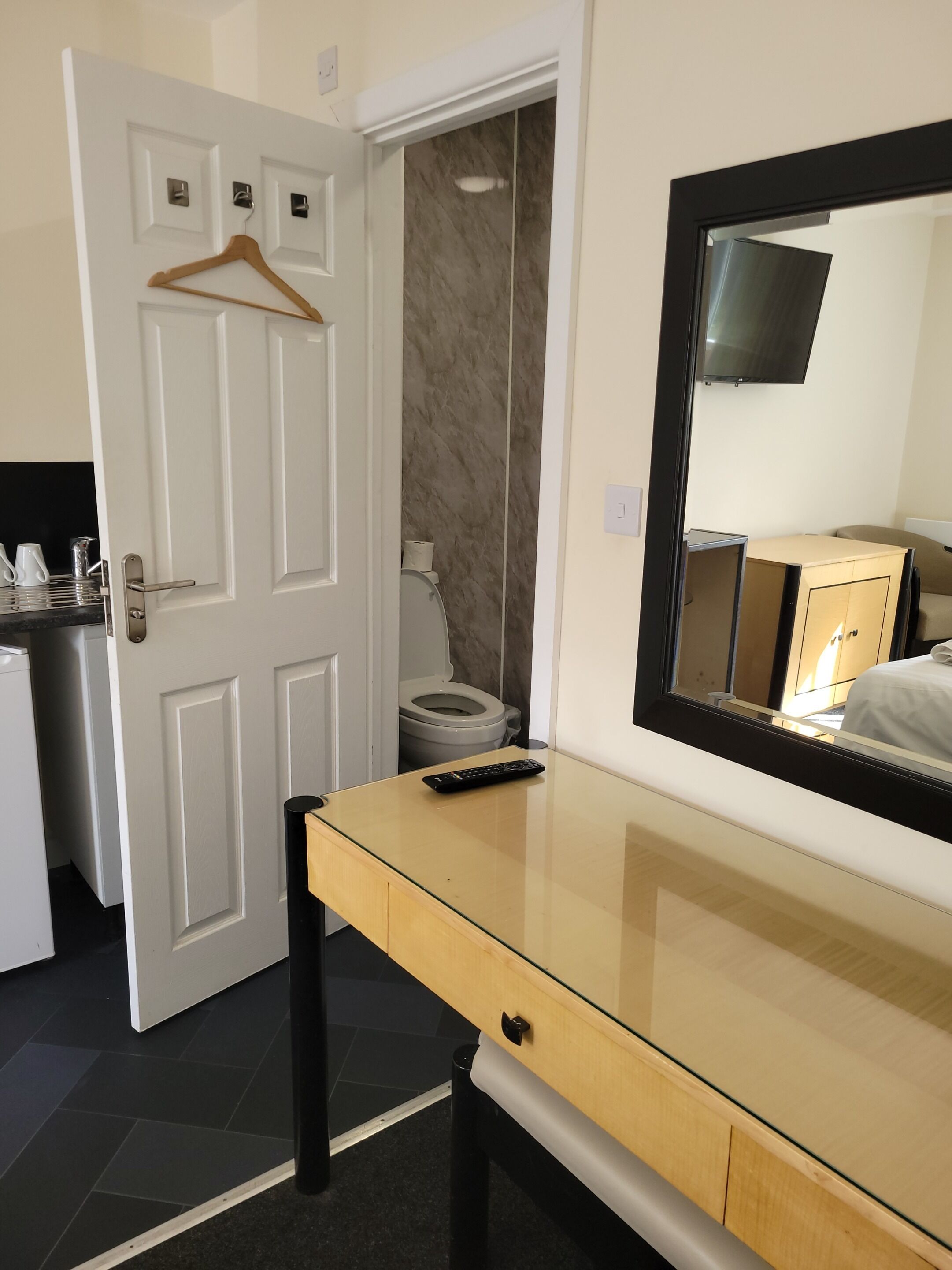 A hotel room with a wooden desk, a mirror, a television, a toilet, and a small kitchenette.