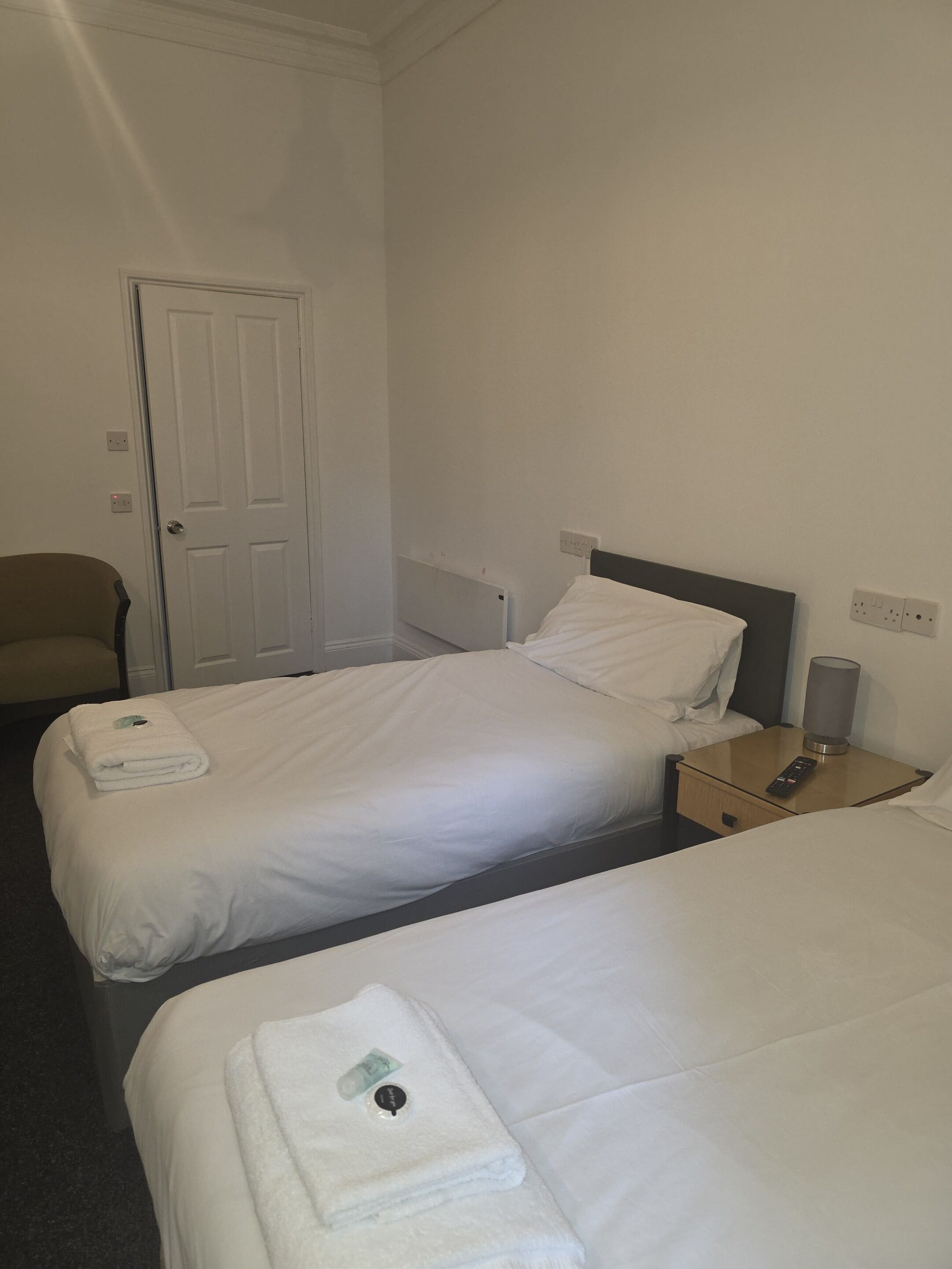Habitació d'hotel amb dos llits individuals, una cadira, una tauleta de nit amb una làmpada i una porta.