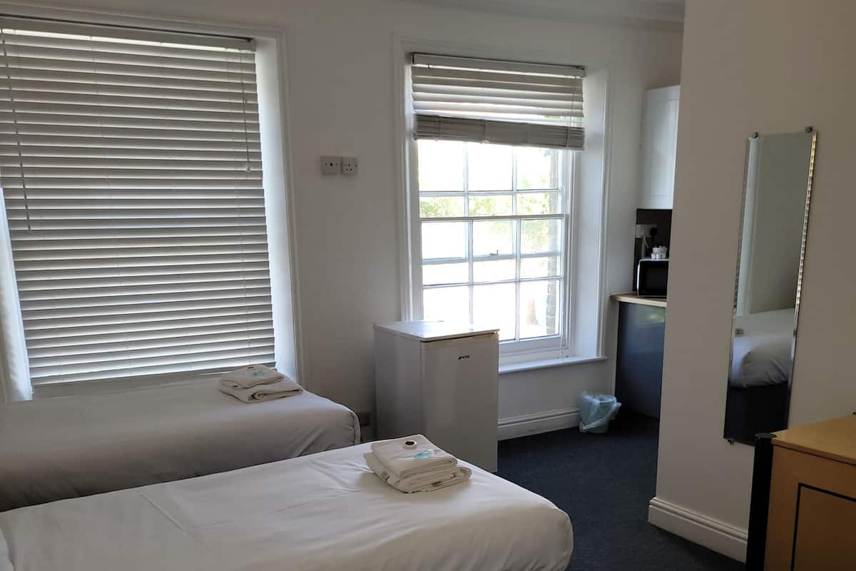 Une chambre d’hôtel avec deux lits, une petite kitchenette, un miroir et une fenêtre avec des stores.