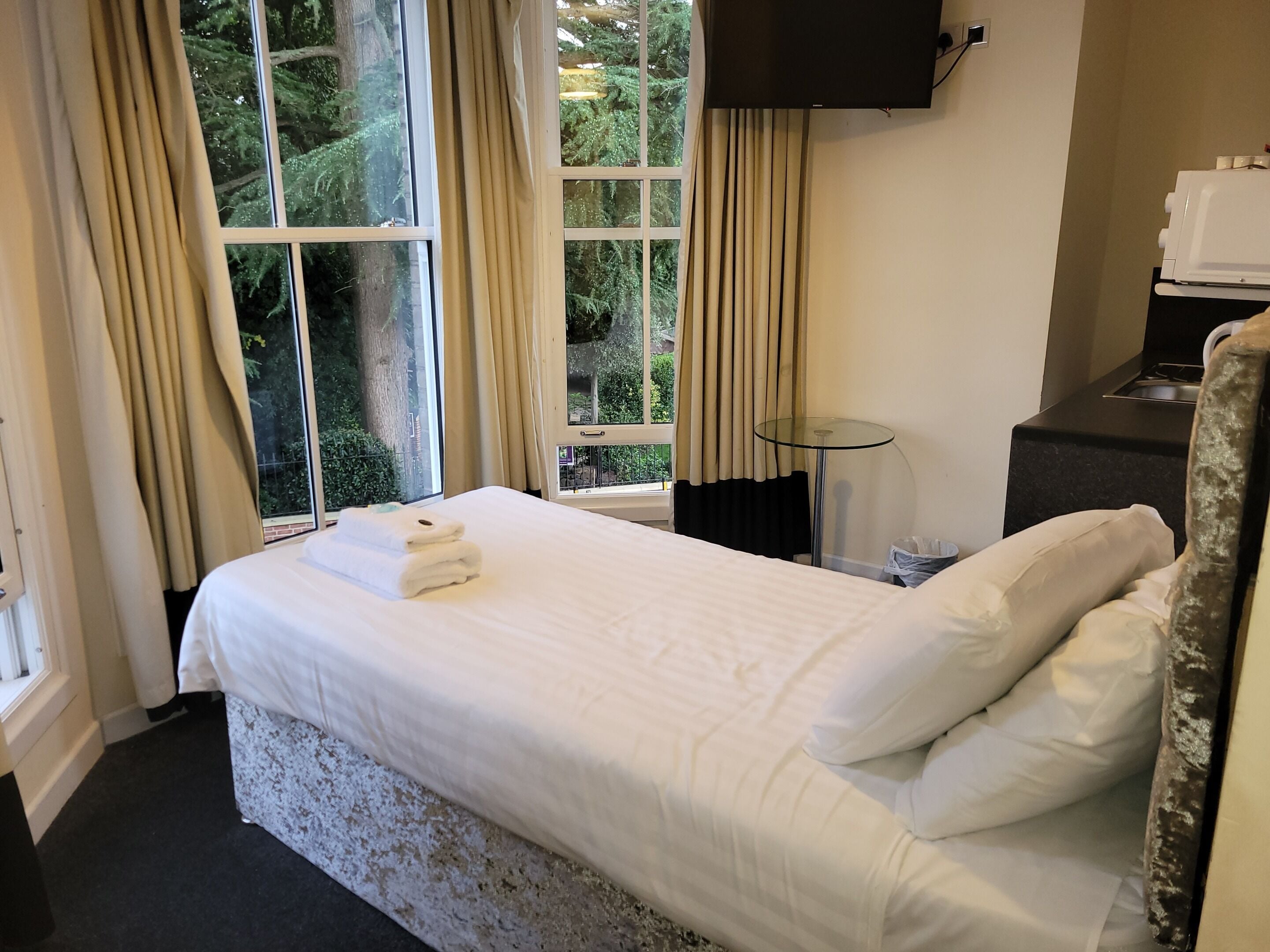 A neatly made bed with white linens and a folded towel, a small round table, a flat-screen TV mounted on the wall, and a window with curtains.