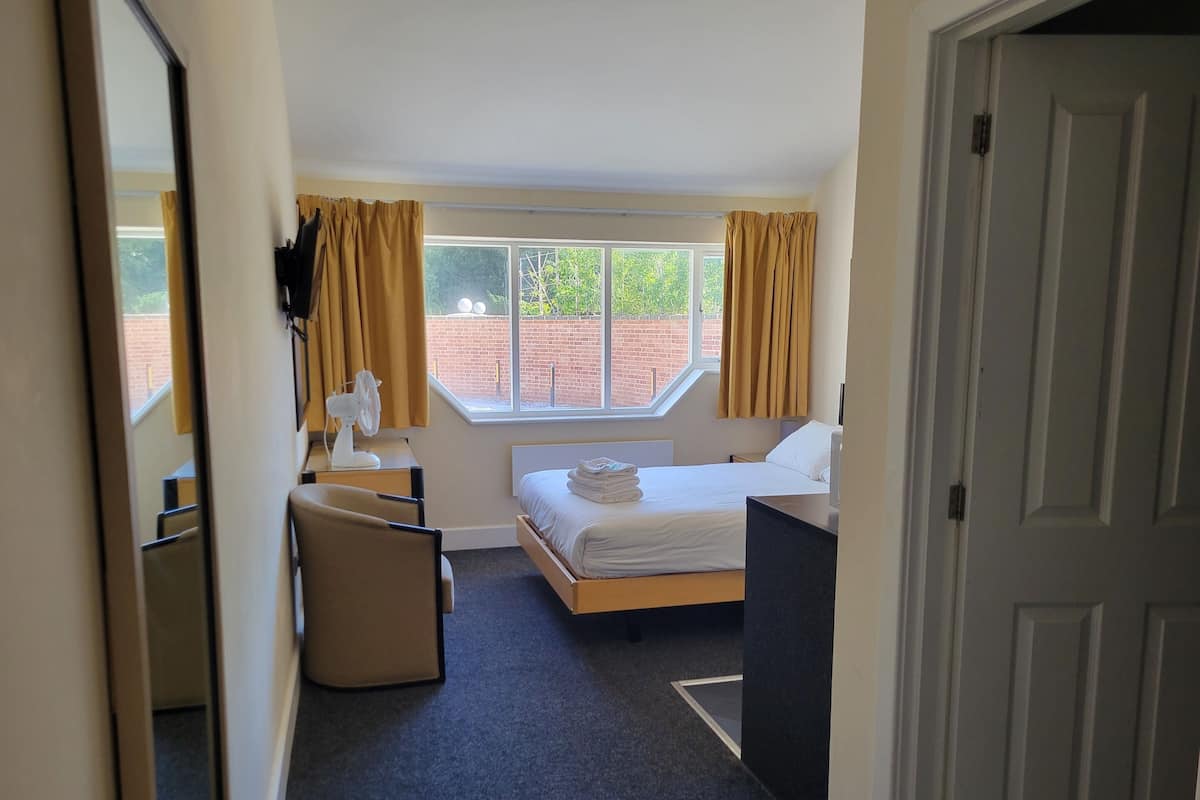 Une chambre d’hôtel avec un lit, un bureau, une chaise, une fenêtre avec des rideaux et une vue sur un mur de briques et de la verdure à l’extérieur.