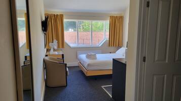 Quarto de hotel com cama, escrivaninha, cadeira, janela com cortinas e vista para uma parede de tijolos e vegetação lá fora.