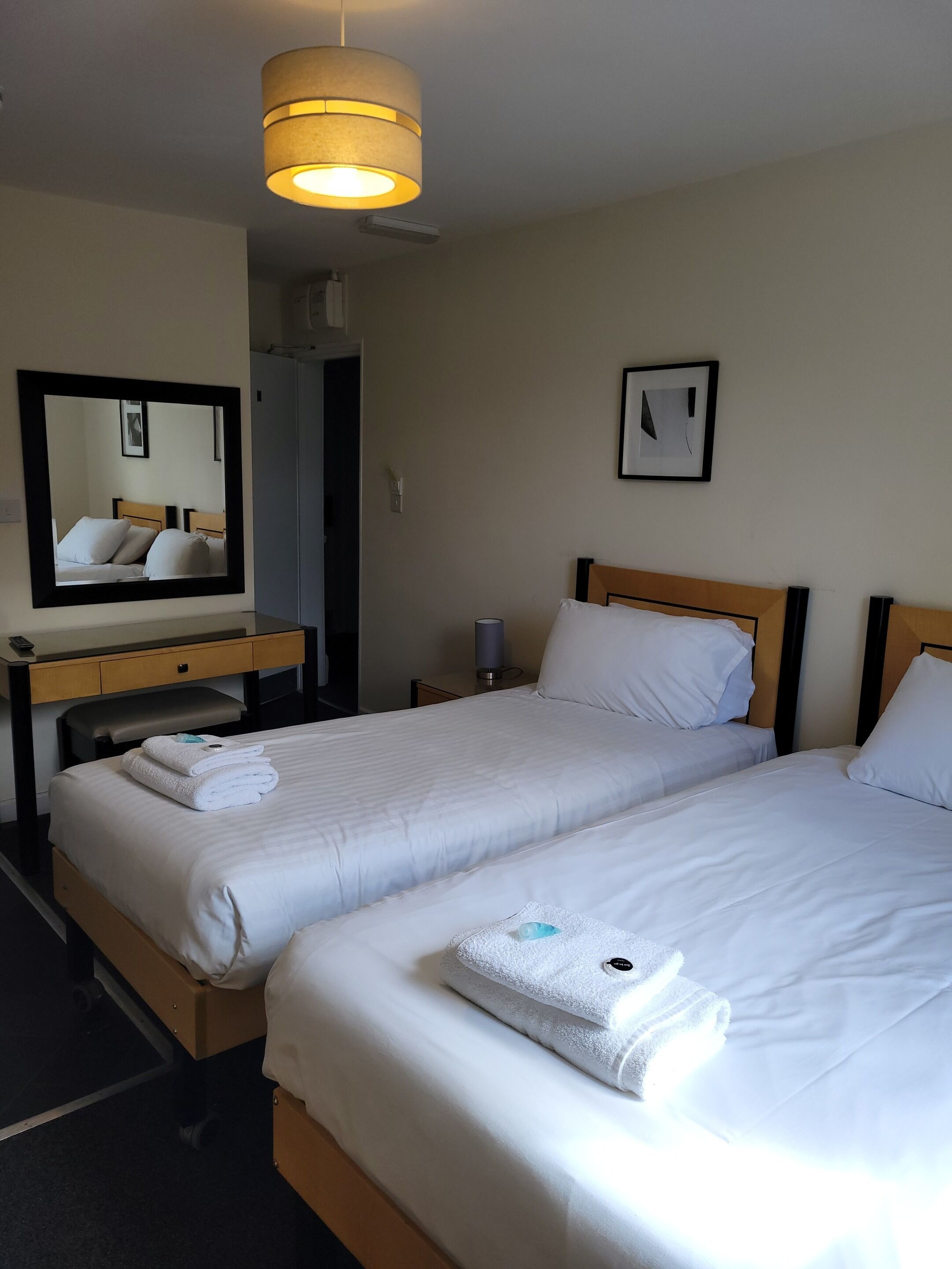 A hotel room with two single beds, a desk with a chair, a mirror, a bedside table with a lamp, and a framed picture on the wall.