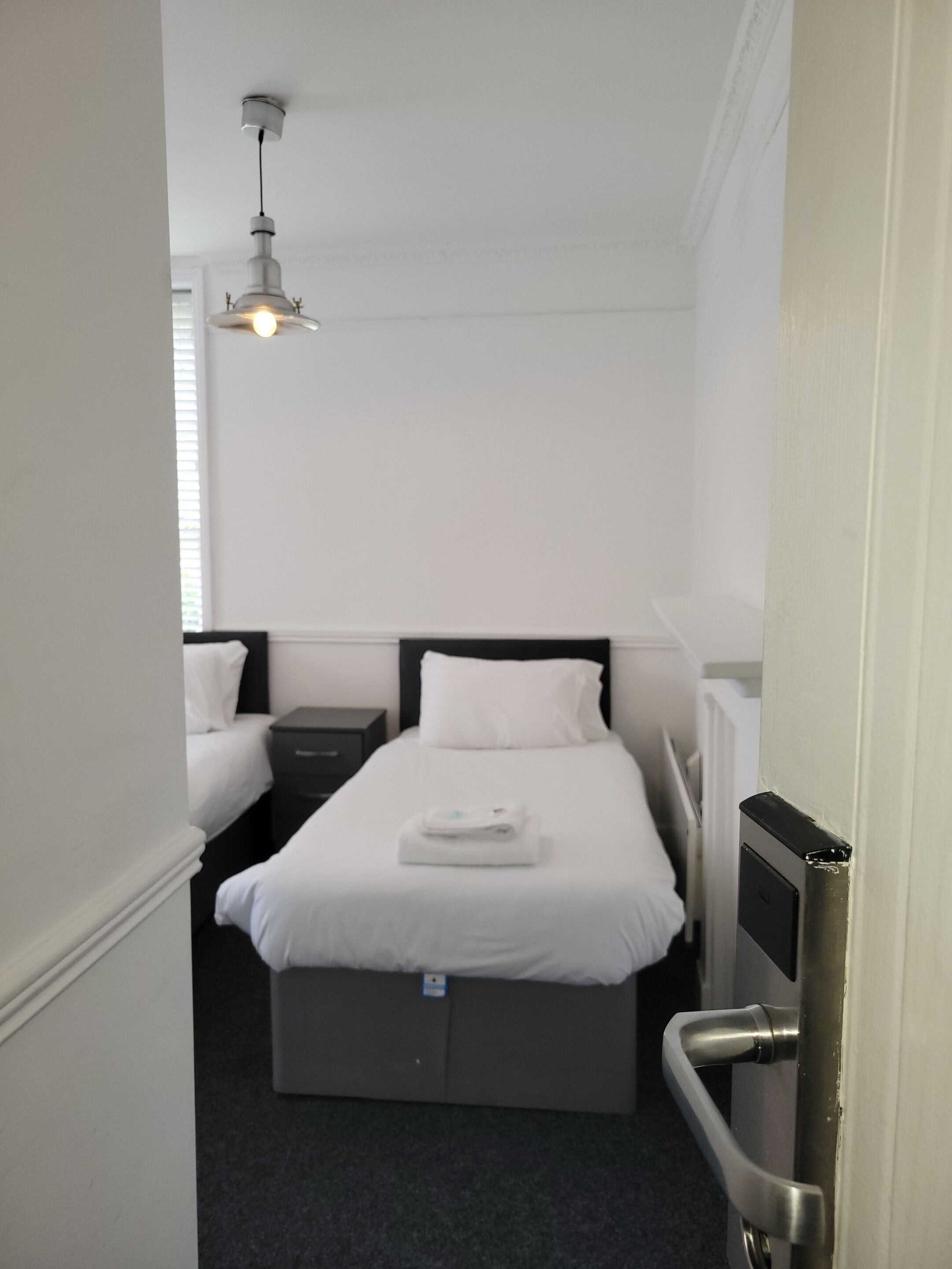 A small, neatly arranged hotel room with a single bed, a bedside table, and a hanging light fixture.
