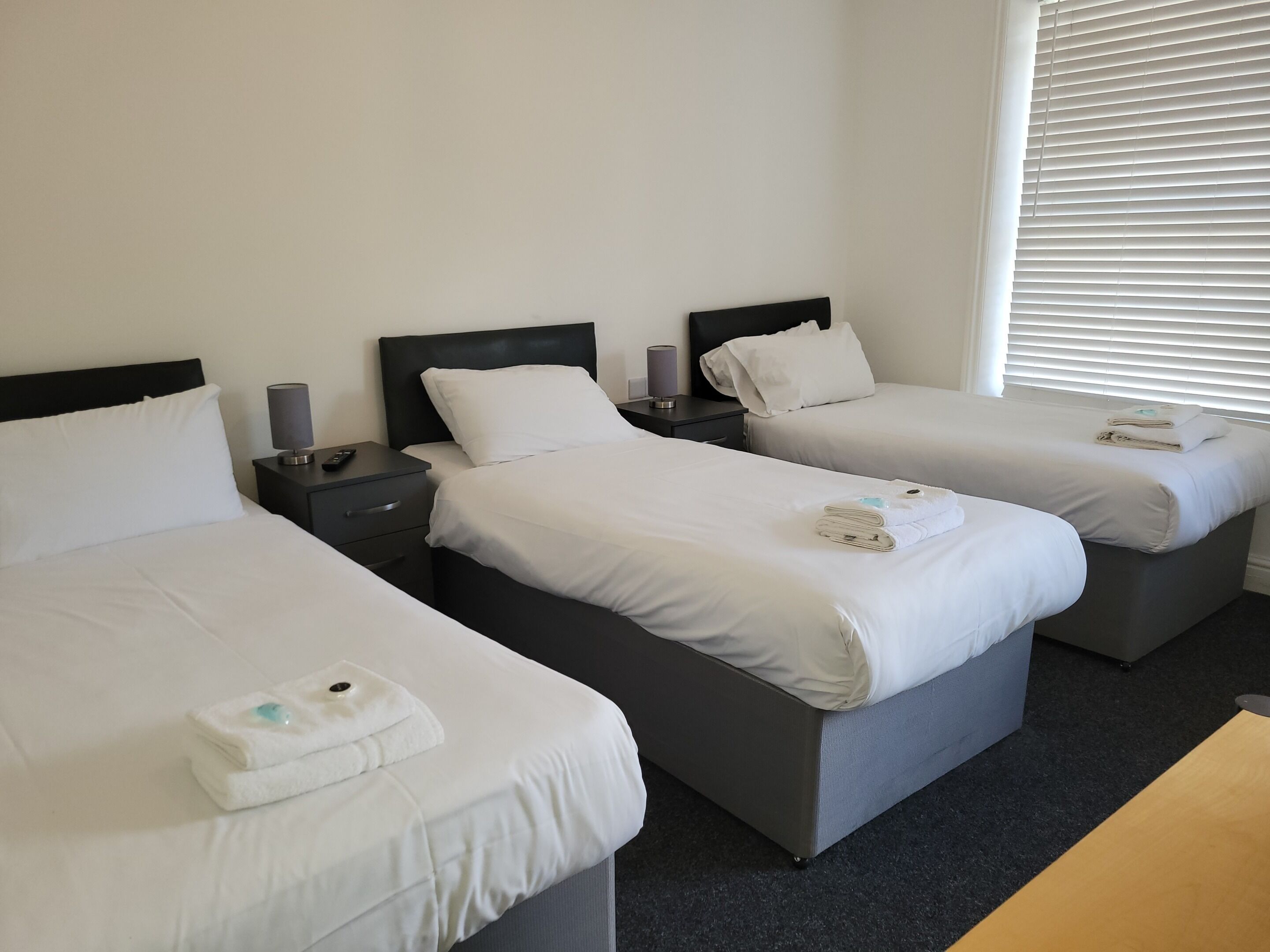 A hotel room with three single beds, each with white linens and pillows, a bedside table with a lamp, and a window with blinds.