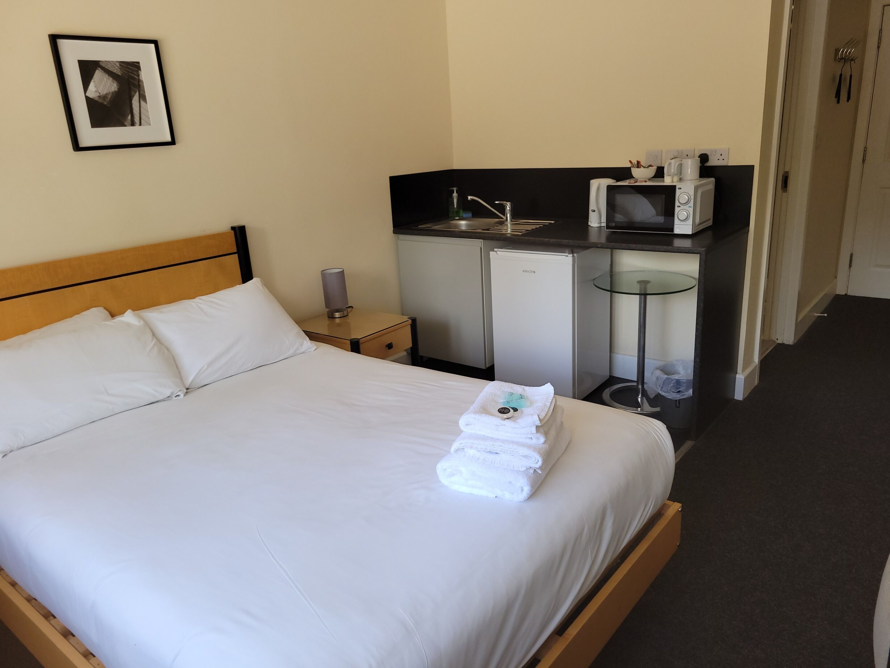 A hotel room with a bed, a small kitchenette, a refrigerator, and a glass table.