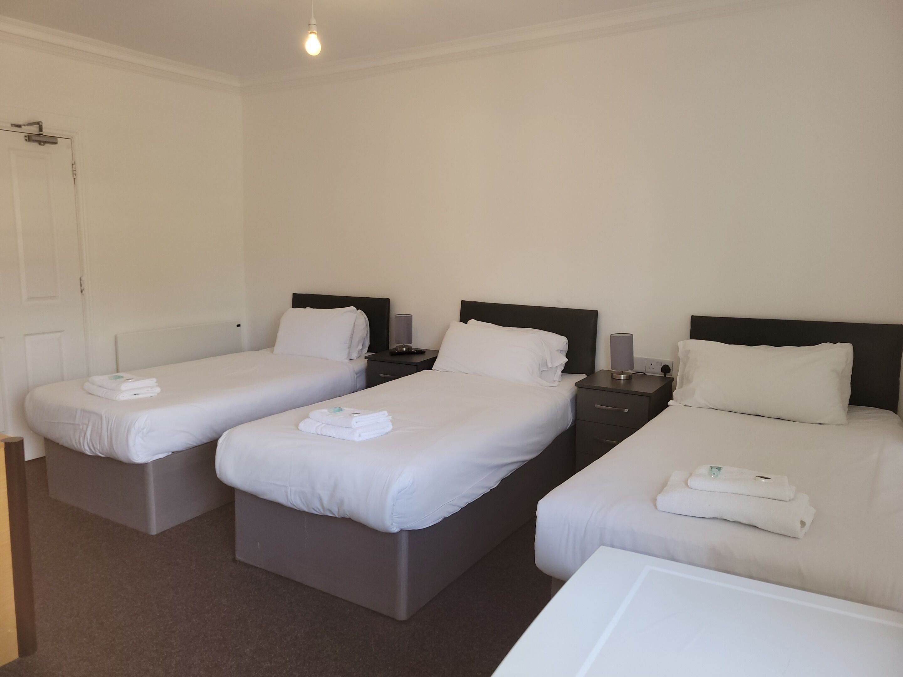 A hotel room with three single beds, each with a bedside table and lamp.