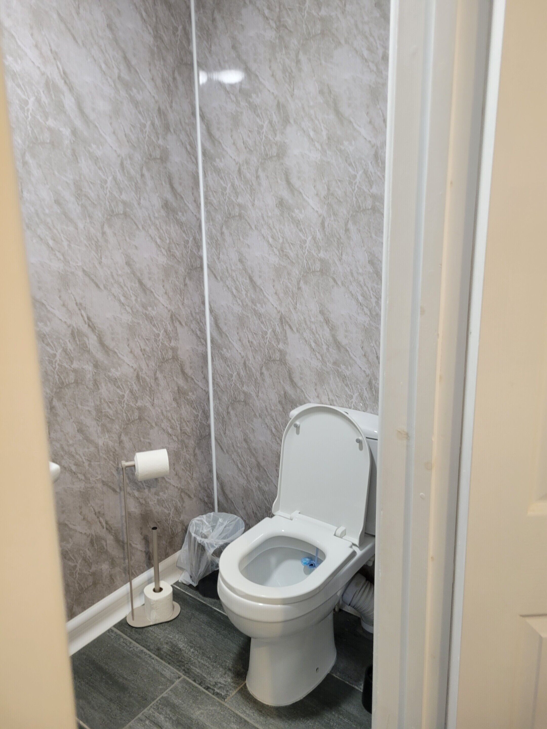 A bathroom with a toilet, marble walls, a trash bin, and a floor lamp.