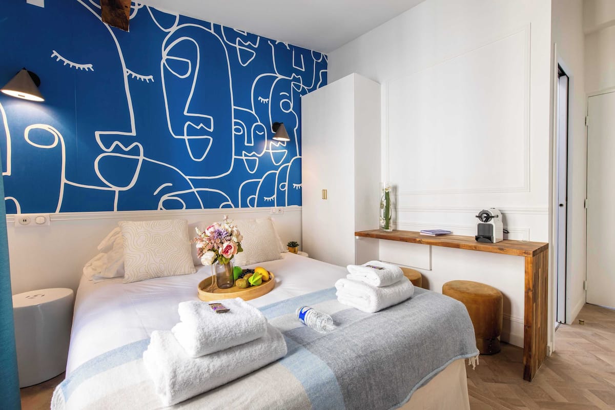 Quarto de hotel moderno com uma cama grande, uma mesinha de cabeceira com um vaso de flores e um banco de madeira.