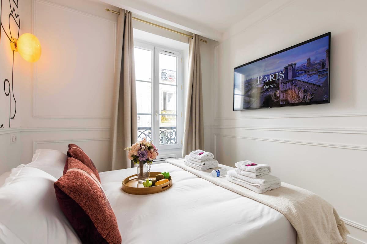 Quarto de hotel com uma cama, uma TV exibindo Paris, uma janela com cortinas e um suporte com artigos de higiene pessoal.