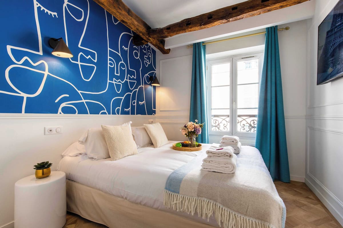 Quarto com uma cama grande, um mural na parede, uma janela com cortinas e uma pequena mesa de cabeceira com uma planta.