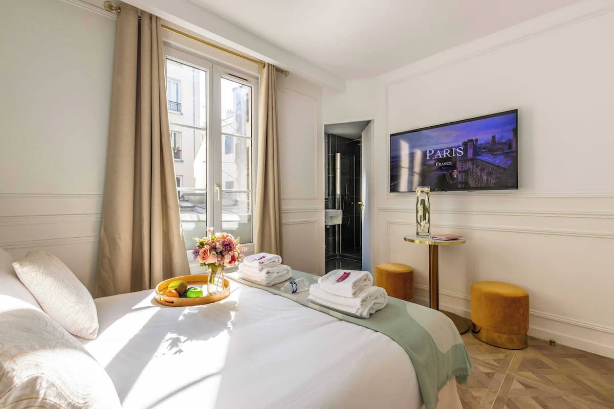 Quarto de hotel com cama, janela com cortinas, televisão na parede e uma mesa pequena com toalhas e uma cesta de frutas.