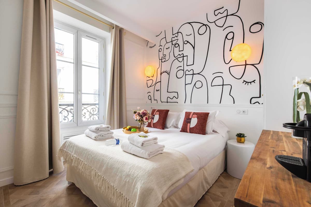 Quarto de hotel com uma cama grande, uma mesa de cabeceira de madeira, uma pequena mesa redonda e uma obra de arte na parede.