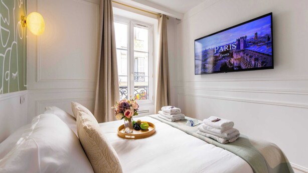 Una habitación de hotel con una cama, un televisor mostrando una escena de París, una ventana con cortinas y una bandeja con toallas y un jarrón de flores.