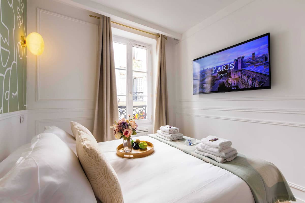 Quarto de hotel com cama, televisão exibindo uma cena de Paris, janela com cortinas e bandeja com toalhas e um vaso de flores.